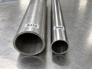 Two stainless steel pipe and tube samples, one marked A312 and one marked A269, lying side by side on a bench